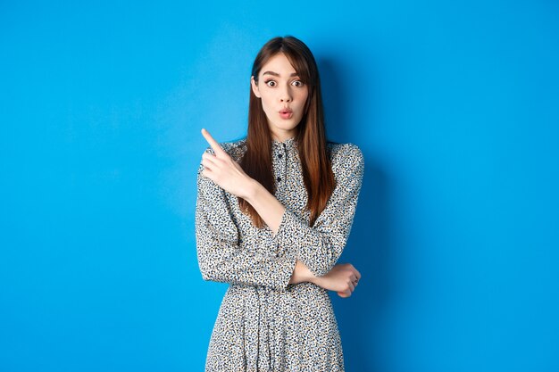 Une jeune femme excitée dit wow, l'air impressionnée et pointant vers la gauche le logo, montrant des nouvelles géniales, debout une jolie robe sur fond bleu