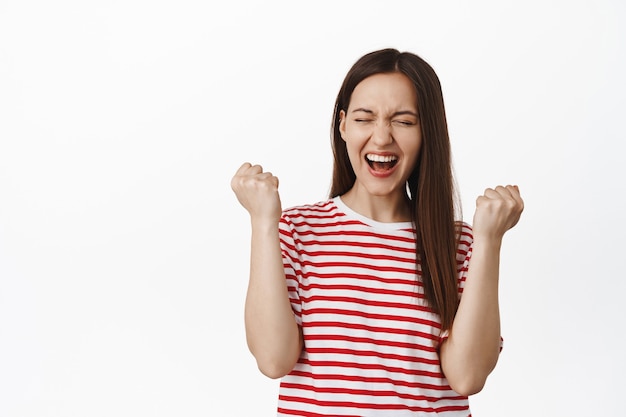 Jeune femme excitée pompe à poing, femme criant oui et triomphant, atteindre l'objectif, célébrer la victoire, debout en t-shirt contre le mur blanc