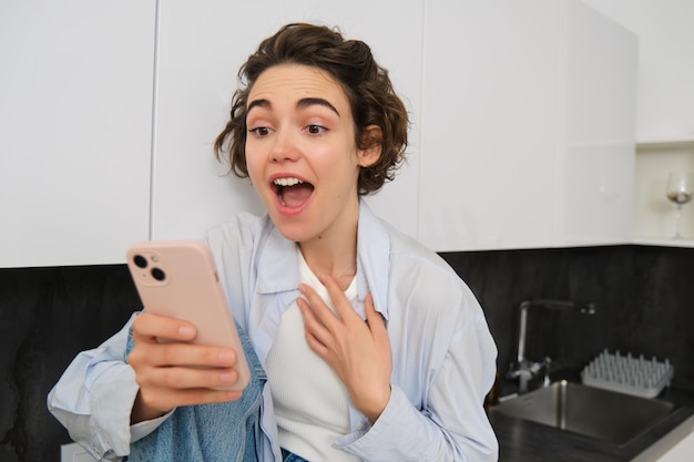 Une jeune femme excitée qui regarde étonnée les conversations sur l'écran du téléphone portable avec quelqu'un voit des nouvelles étonnantes sur sm