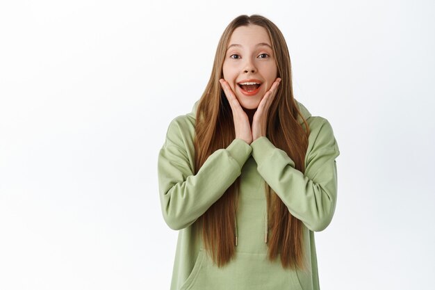 Une jeune femme excitée et surprise étant touchée et émerveillée, se tenant la main sur le visage et souriante étonnée, regarde avec admiration, debout en sweat à capuche sur fond blanc.