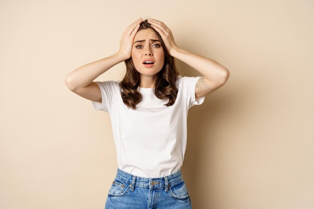 Jeune femme face à une catastrophe, l'air anxieuse dans la panique, tenant les mains sur la tête frustrée, debout sur fond beige.