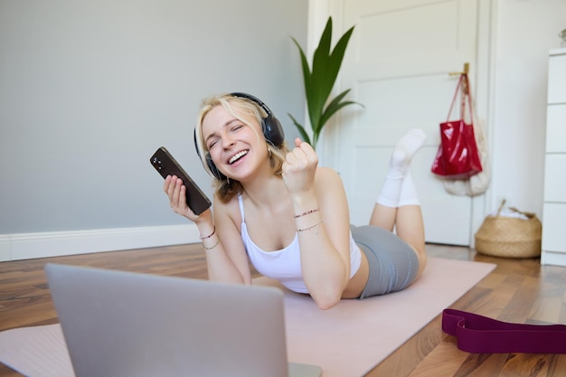 Photo gratuite une jeune femme faisant de l'entraînement physique à la maison allongée sur un tapis de yoga en caoutchouc tenant un smartphone.