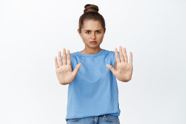 Jeune femme faisant des gestes d'arrêt, disant non, tendant les mains pour interdire quelque chose de mal, debout sur blanc