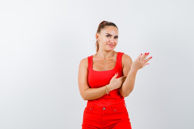 Jeune femme faisant semblant de montrer quelque chose en maillot rouge, pantalon rouge et à la gaieté. vue de face.