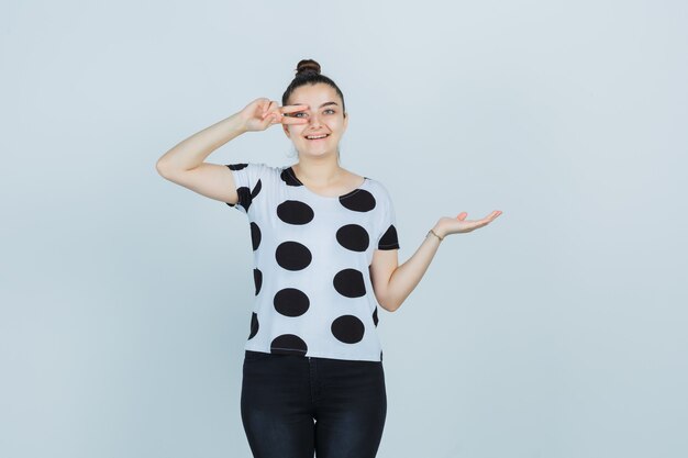 Jeune femme faisant semblant de montrer quelque chose tout en montrant le signe de la victoire en t-shirt, jeans et à la chance. vue de face.