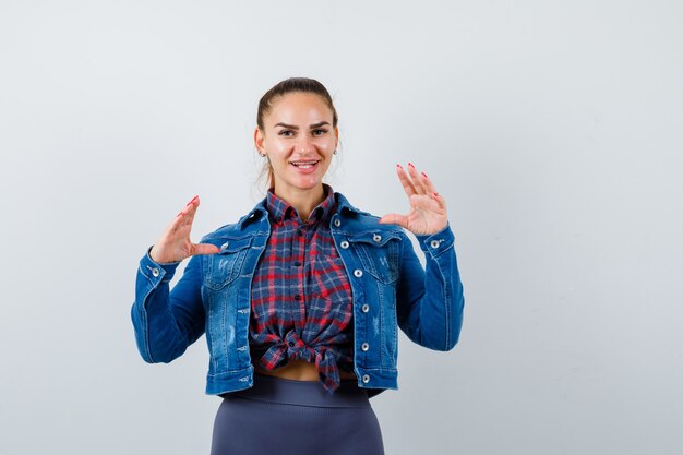 Jeune femme faisant semblant de tenir quelque chose en chemise à carreaux, veste en jean et semblant attrayante, vue de face.