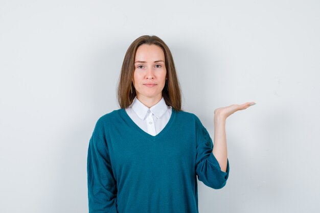 Jeune femme faisant semblant de tenir quelque chose dans un pull sur une chemise blanche et ayant l'air confiant, vue de face.