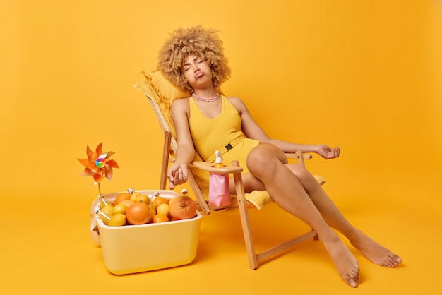 Photo gratuite jeune femme fait la sieste et prend un bain de soleil pendant la journée d'été pose sur une chaise longue vêtue d'un maillot de bain a des jambes minces mange des fruits tropicaux frais et boit des boissons du réfrigérateur portable concept de vacances