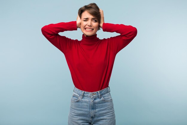 Jeune femme fatiguée agacée en pull rouge et jeans fermant malheureusement les oreilles avec les mains sur fond bleu isolé