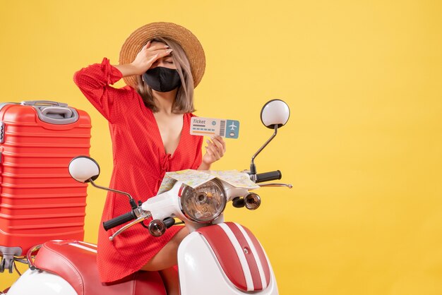 Jeune femme fatiguée sur un cyclomoteur avec une valise rouge tenant un ticket mettant la main sur sa tête