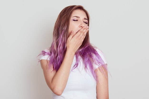 Une jeune femme fatiguée endormie s'habille de t-shirt décontracté blanc bâillant, couvrant la bouche ouverte avec la paume, garde la bouche fermée, a besoin de dormir plus, posant isolée sur un mur gris.