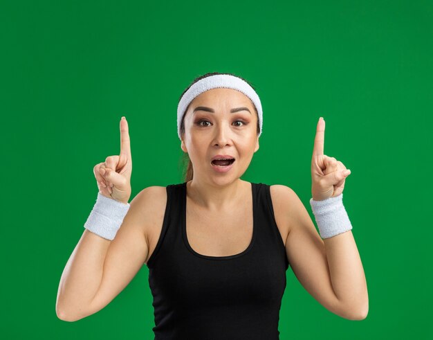 Jeune femme fitness avec bandeau et brassards surpris montrant des index debout sur un mur vert