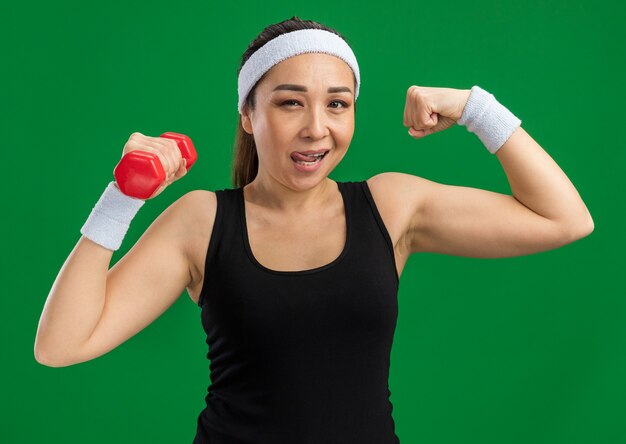 Jeune femme fitness avec bandeau avec haltères faisant des exercices levant le poing montrant des biceps souriant confiant debout sur un mur vert