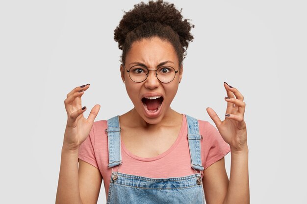 Une jeune femme folle à la peau sombre et furieuse fait des gestes de colère, hurle de stress, porte des vêtements décontractés, se tient contre un mur blanc. La femme afro-américaine exprime sa rage, a une mauvaise expression.