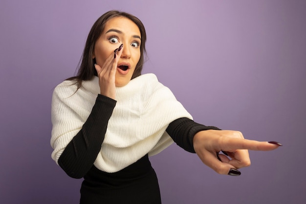 Photo gratuite jeune femme avec un foulard blanc à la surprise en pointant avec l'index à quelque chose qui raconte un secret avec la bouche nera main