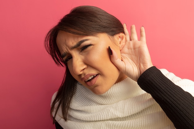 Photo gratuite jeune femme avec un foulard blanc tenant la main près de l'oreille en essayant d'écouter les potins debout sur le mur rose