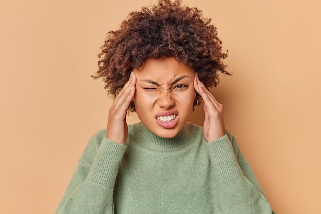 Photo gratuite une jeune femme frisée frustrée souffre de migraine garde les mains sur les tempes a des maux de tête serre les dents pour révéler la douleur porte un pull décontracté isolé sur fond beige. notion de problèmes de santé