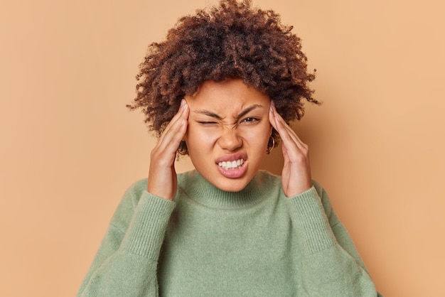 Une jeune femme frisée frustrée souffre de migraine garde les mains sur les tempes a des maux de tête serre les dents pour révéler la douleur porte un pull décontracté isolé sur fond beige. Notion de problèmes de santé