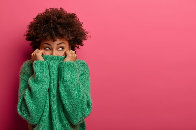 Une jeune femme frisée positive tire un pull vert sur la tête, s'amuse et cache le visage, regarde par le dessous, disparaît dans ses vêtements, regarde avec plaisir, isolé sur un mur rose, espace copie