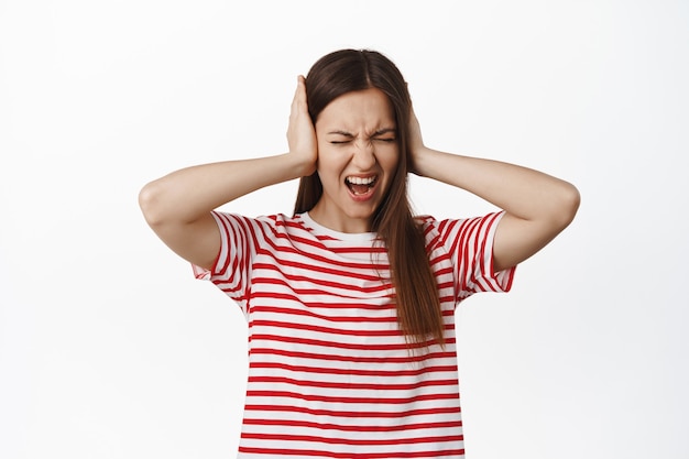 Jeune femme frustrée et agacée criant dans le déni, refusant d'écouter, marre du bruit fort agaçant, couvre les oreilles avec les mains et crie, debout sur un mur blanc