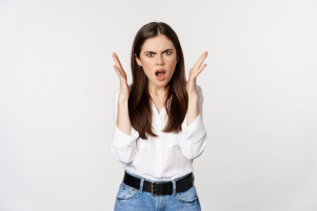 Jeune femme frustrée et en colère haletant, l'air choquée et contrariée par la caméra, offensée par qch, debout gênée sur fond blanc.