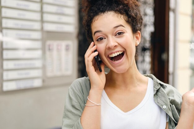 Jeune femme gaie aux cheveux bouclés foncés en chemise kaki et t-shirt blanc parlant au téléphone portable et regardant joyeusement à huis clos avec la bouche ouverte sur la rue de la ville