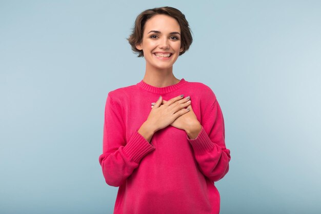 Jeune femme gaie aux cheveux courts foncés en pull rose tenant les mains sur la poitrine tout en regardant joyeusement à huis clos sur fond bleu