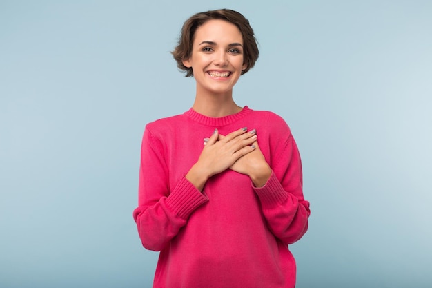 Jeune femme gaie aux cheveux courts foncés en pull rose tenant les mains sur la poitrine tout en regardant joyeusement à huis clos sur fond bleu