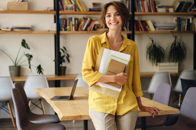 Jeune femme gaie en chemise jaune appuyée sur le bureau avec bloc-notes et papiers à la main tout en regardant joyeusement à huis clos dans un bureau moderne