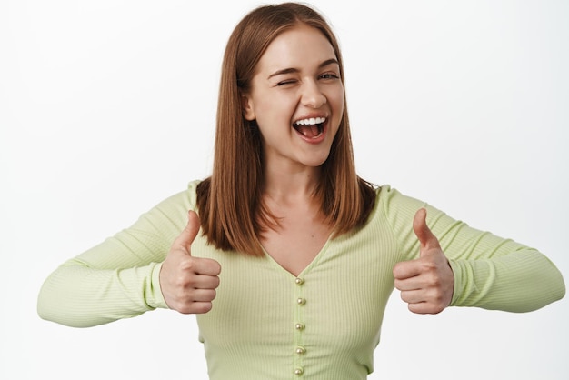 Photo gratuite jeune femme gaie faisant un clin d'œil et montrant son soutien avec les pouces vers le haut en souriant donne un coup de pouce à la confiance louant le beau travail compliment et recommande le fond blanc de l'entreprise