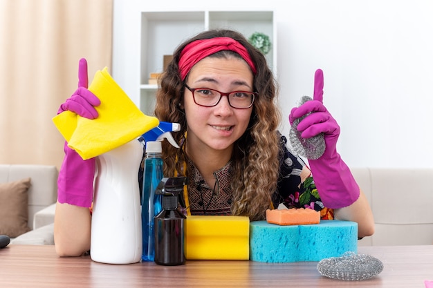 Jeune femme en gants de caoutchouc regardant la caméra en souriant montrant les index assis à la table avec des produits de nettoyage et des outils dans la salle de séjour lumineuse