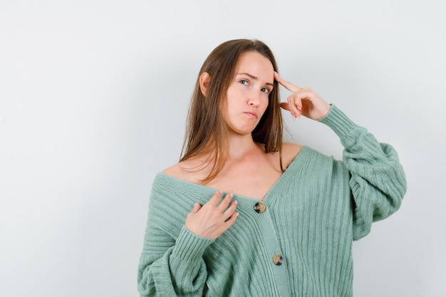 Jeune femme en gardant le doigt sur le front en cardigan en laine et à l'hésitation. vue de face.