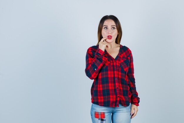 Jeune femme gardant le doigt près de la bouche dans une chemise à carreaux, un jean et l'air pensive. vue de face.