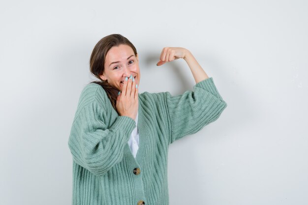 Jeune femme gardant la main sur la bouche, montrant les muscles du bras en blouse, cardigan et l'air joyeux. vue de face.