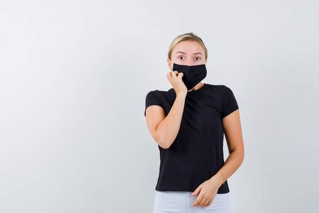 Jeune femme gardant la main sur la joue en t-shirt noir, masque et à la vue étonnée, de face.