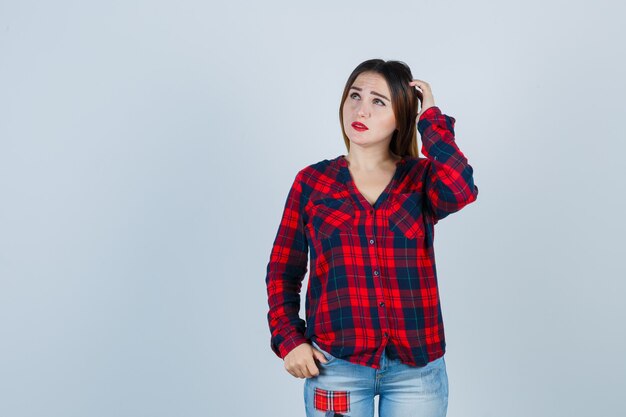 Jeune femme gardant la main sur la tête tout en regardant une chemise à carreaux, un jean et l'air réfléchi. vue de face.