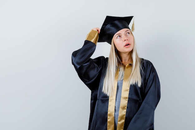 Jeune femme gardant la main sur la tête en uniforme de diplômé et l'air pensive.