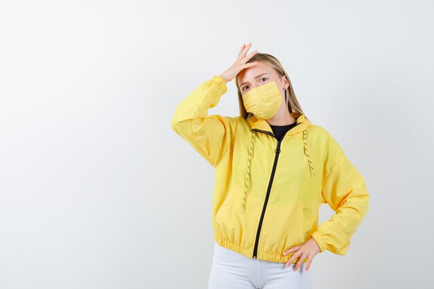 Jeune femme gardant la main sur la tête en veste, pantalon, masque et regardant pensif, vue de face.