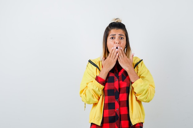 Jeune femme gardant les mains sur la bouche en chemise à carreaux, veste et choquée. vue de face.