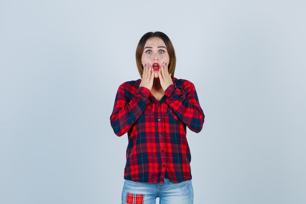 Jeune femme gardant les mains sur les joues en chemise à carreaux, jeans et l'air terrifiée, vue de face.