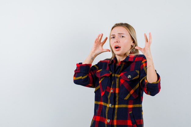 Jeune femme gardant les mains de manière agressive en chemise à carreaux et à la furieuse, vue de face.