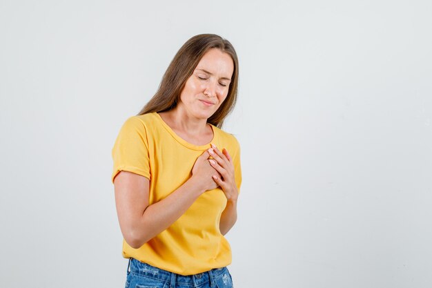 Jeune femme gardant les mains sur la poitrine avec les yeux fermés en t-shirt, short et à la reconnaissance. vue de face.