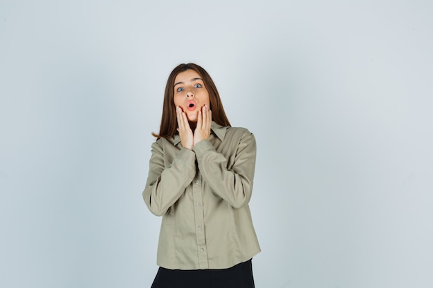 Jeune femme gardant les mains près de la bouche ouverte en chemise, jupe et semblant étonnée. vue de face.