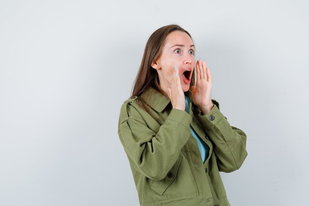 Jeune femme gardant les mains près de la bouche ouverte en veste verte et se demandant. vue de face.