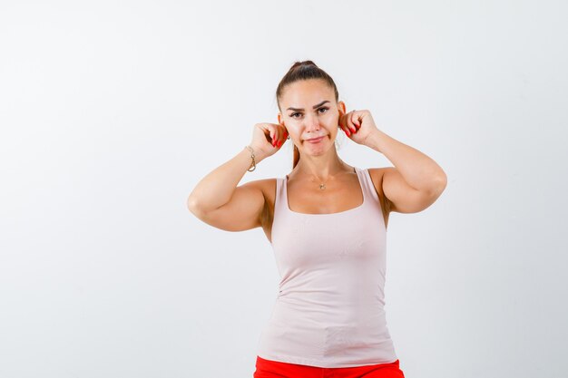 Jeune femme gardant les oreilles avec les mains en débardeur beige et à la recherche de mignon. vue de face.