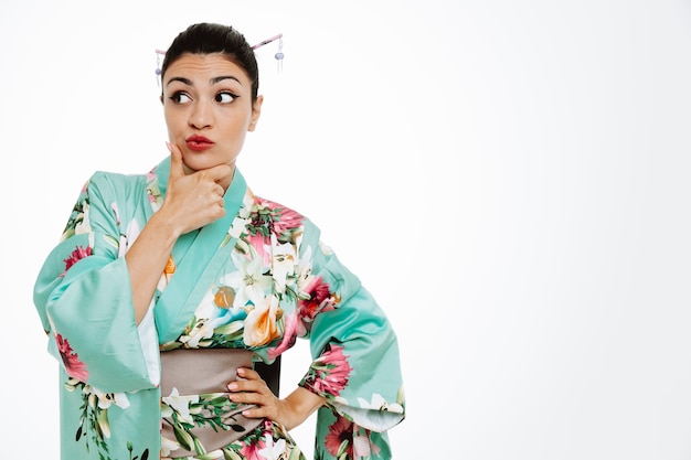 jeune femme geisha en kimono japonais traditionnel regardant de côté avec une expression pensive avec la main sur son menton pensant debout sur un mur blanc