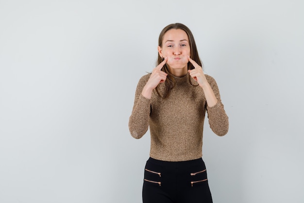 Photo gratuite jeune femme gonflant les joues et mettant l'index sur les joues en pull doré et pantalon noir et à la recherche amusée. vue de face.