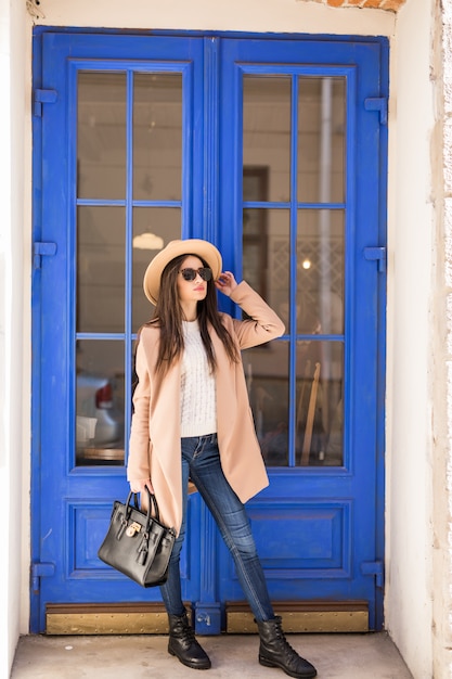 Jeune femme habillée décontractée en chapeau de manteau brillant et lunettes de soleil se tient devant la porte bleue