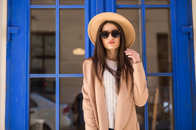 Photo gratuite jeune femme habillée décontractée en chapeau de manteau brillant et lunettes de soleil se tient devant la porte bleue