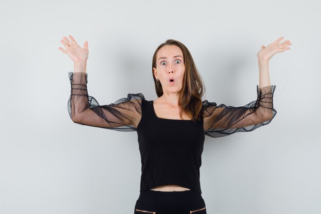 Jeune femme haussant les épaules en chemisier noir et pantalon noir et à la vue choquée, de face.